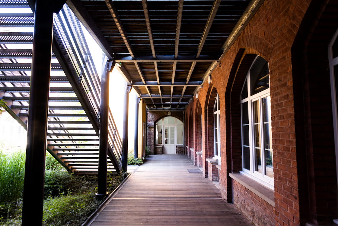 Allée extérieure en bois sous des arcades qui donnent accès à un escalier en métal sur la gauche et des salles de classe sur la droite.