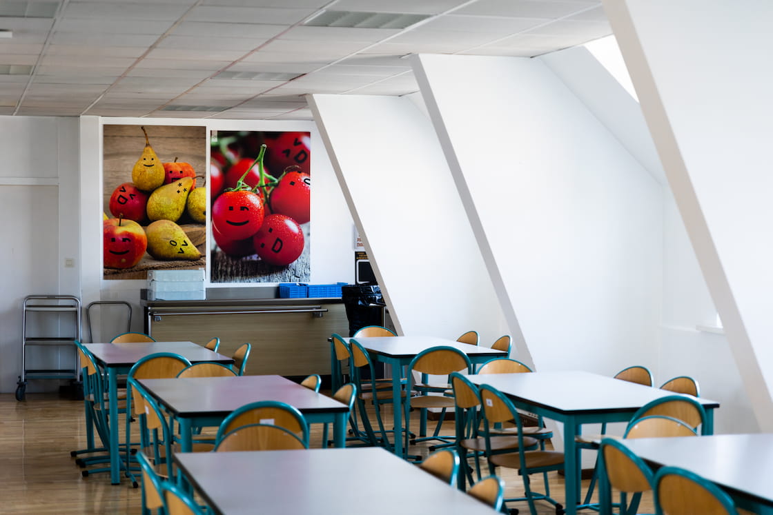 Des tables du restaurant sont agencées près des fenêtres et des photos de pommes, poires et tomates avec des smileys dessus sont accrochées au mur.